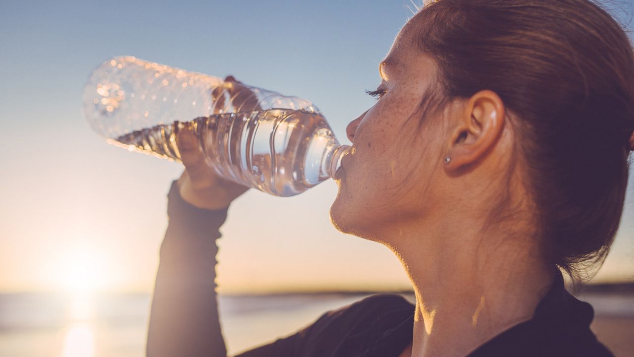 A woman drinks water from a water bottle on the beach while the sun sets in the distance.