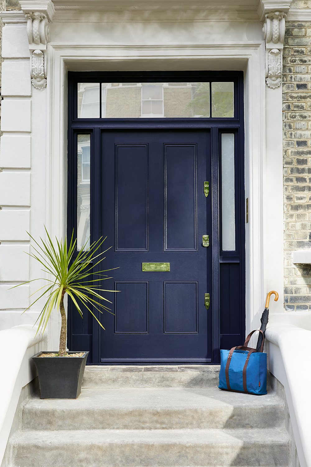 A navy blue front door on an off-white stone staircase and stoop. There is a plant in a black pot on the stoop and a blue bag and umbrella.