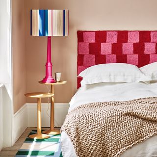 A pink bedroom with a red check headboard and a lamp with a multicoloured lamp shade on the bedside table