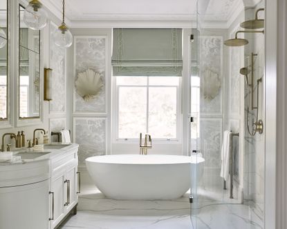 An example of bathroom storage tips showing a white freestanding bath in the middle of a bathroom with a large sash window, a double headed shower and his and hers sinks with white cabinets