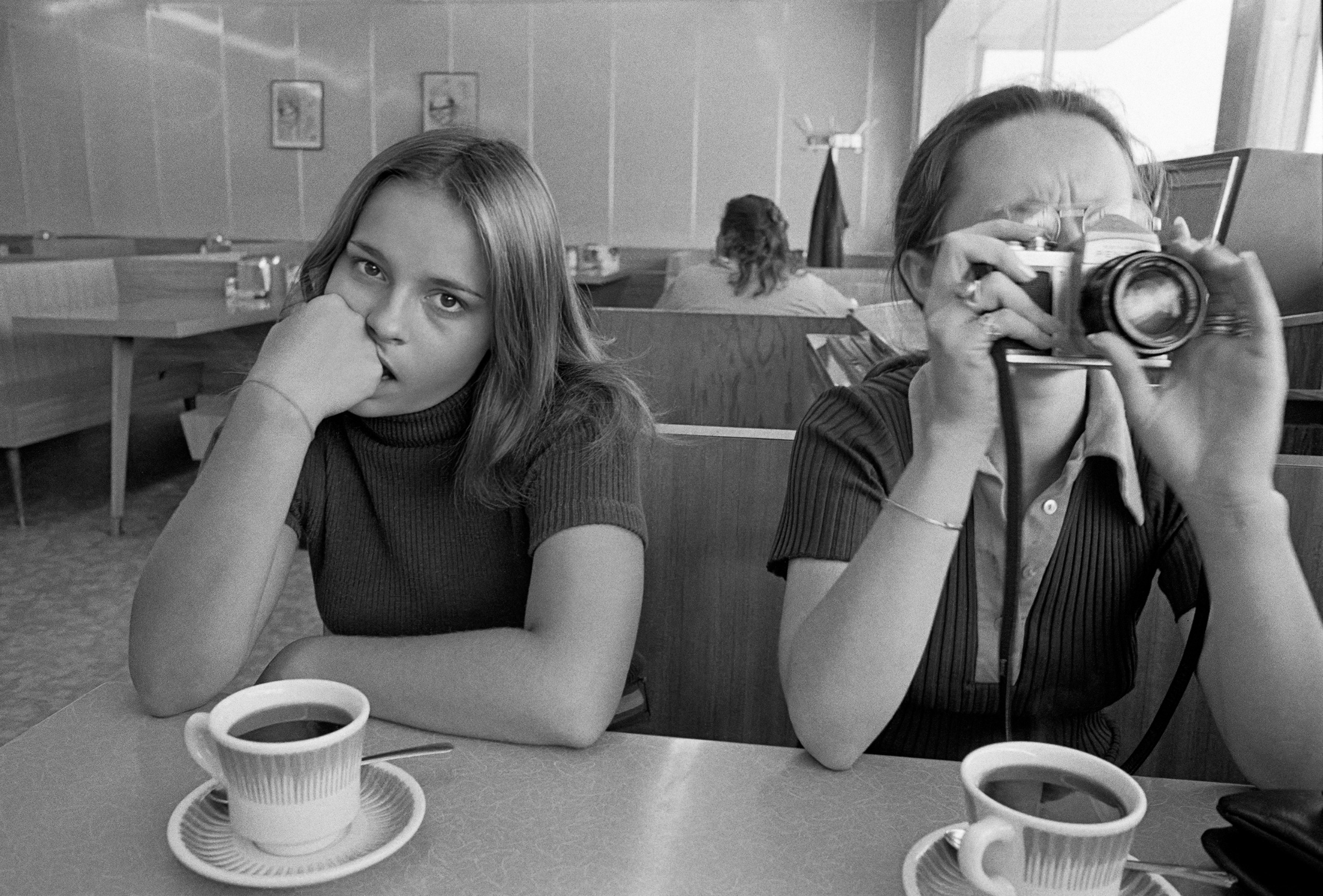 Two young women sit in a diner booth with cups of coffee, one resting her chin on her fist and staring at the camera while the other points a 35mm camera straight back at the photographer.