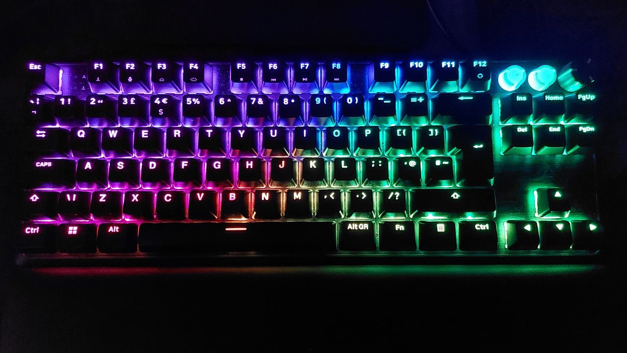 The Corsair K70 Pro TKL gaming keyboard in a dark room, with the RGB lights all powered on via wired connection. All of the keys are lit up in a rainbow of colours.