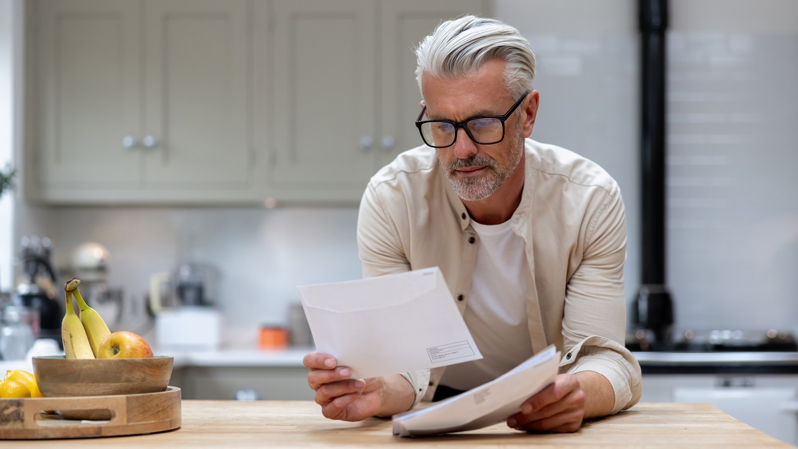 An older man stands at the kitchen counter, looking over his investment statements.