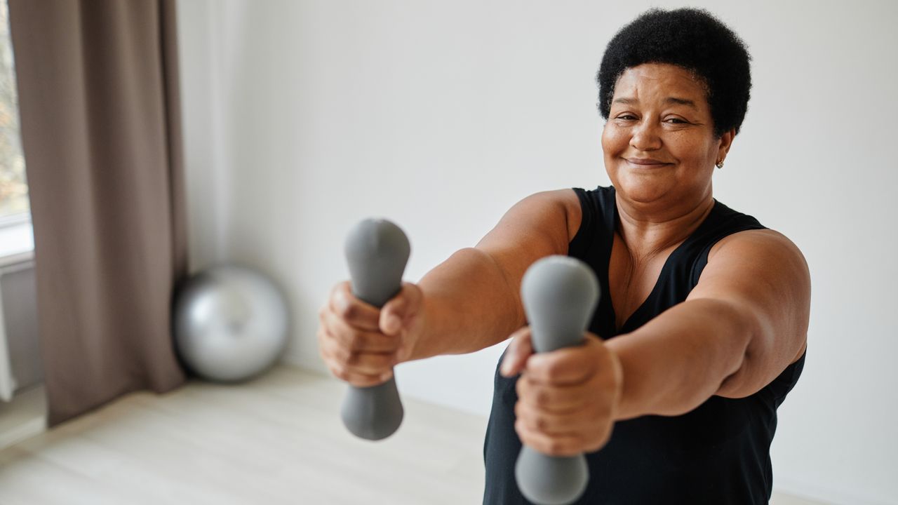 A woman is working out at home in a sparsely decorated room. She wears a sports top and is holding two lightweight dumbbells directly out in front of her shoulders. Behind her we see a window and an exercise ball. 