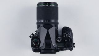 Top view of a black DSLR camera with a Pentax 28-105mm zoom lens attached on a light gray background