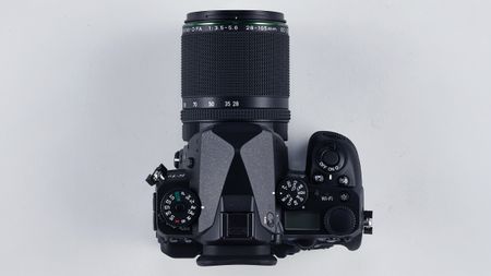 Top view of a black DSLR camera with a Pentax 28-105mm zoom lens attached on a light gray background