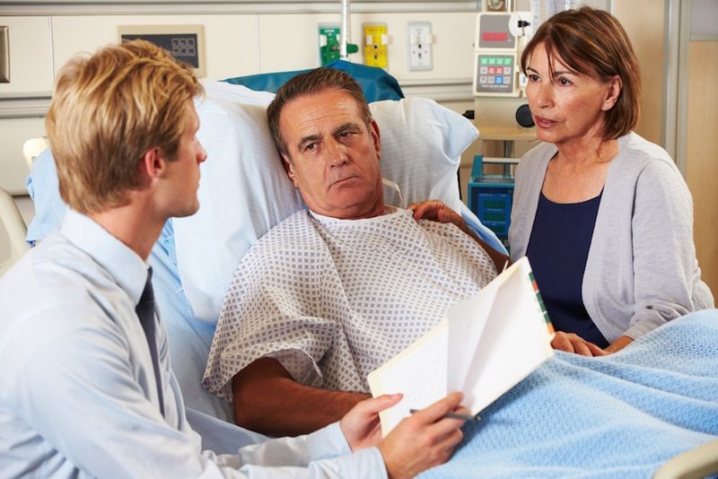 A man lays in a hospital bed and talks with a doctor while his wife looks on.