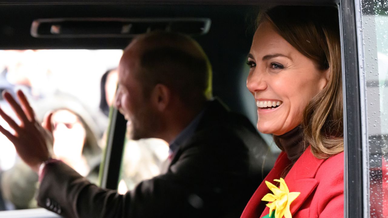 Prince William riding in a car waving to fans out a window sitting next to Princess Kate, wearing a red coat with a yellow daffodil pin and laughing