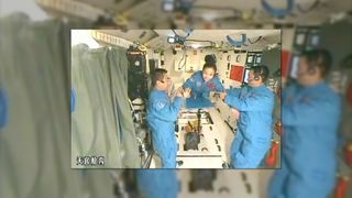 two men and a woman in matching blue coveralls float inside a space lab