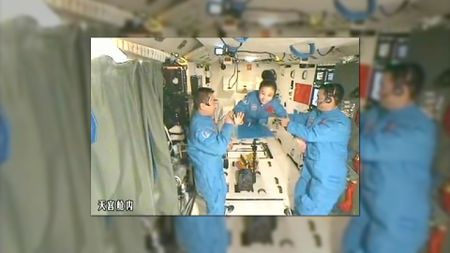 two men and a woman in matching blue coveralls float inside a space lab