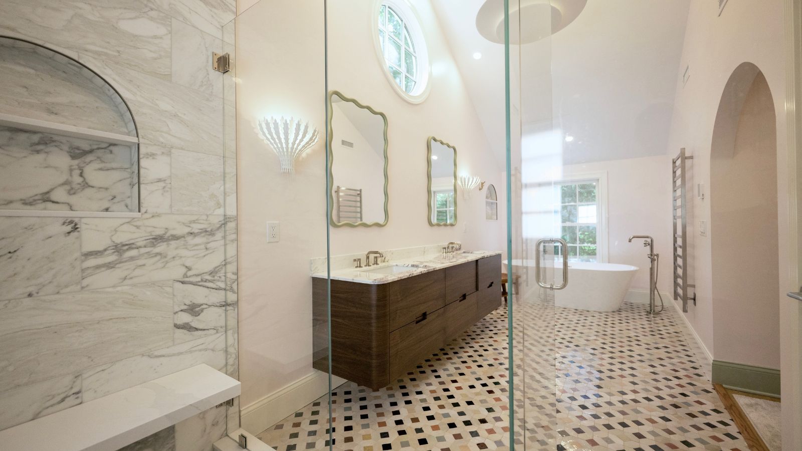 A bright bathroom with a tiled floor, marble shower, wooden cabinetry, white countertops, and scalloped mirrors. In the far end of the bathroom is a white bathtub underneath a window.