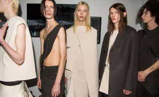 Five models wearing looks from the Rick Owens collection. One model is wearing dark coloured bottoms with a cream piece over the top. Another model is wearing dark brown coloured bottoms and a dark brown piece that exposes the arms and chest. The third model is wearing a cream layered cape style piece. The fourth model is wearing a cream piece with dark grey coat over the top. And the fifth model is wearing a dark brown layered piece with a strap