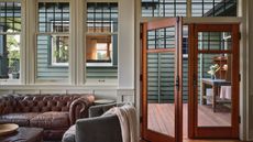 View from inside a home through paneled windows and wooden doors, with brown leather sofa and gray armchair pictured in foreground. Through the window is another building with a green-blue exterior.
