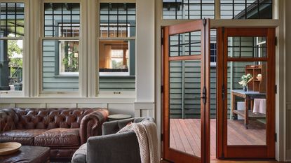 View from inside a home through paneled windows and wooden doors, with brown leather sofa and gray armchair pictured in foreground. Through the window is another building with a green-blue exterior.