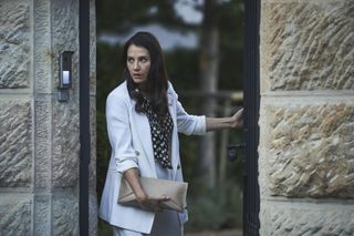 Charlie Murphy as Natalie Varga, exiting her home via strong security gate, looking warily to the side, wearing a white suit over a black blouse with white polka dots and carrying a black leather clutch bag