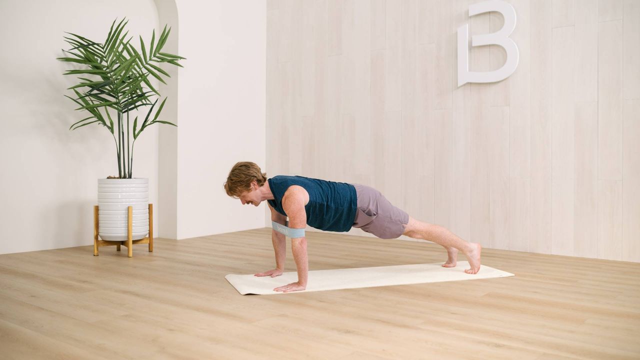 man performing a banded pushup on an exercise mat in a white walled studio corner, wooden floor and a plant behind him on the side.