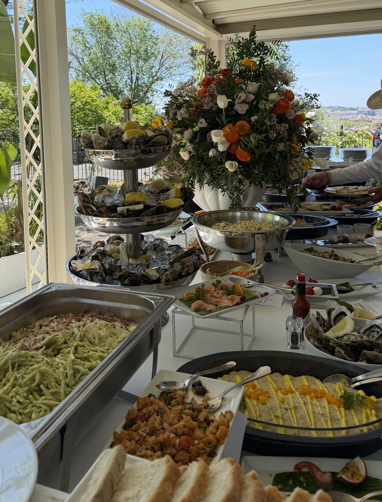 A buffet lunch at Anantara Concorso Roma featuring oysters, seafood, salads and flowers, served in a bright pavilion overlooking gardens and Rome beyond.