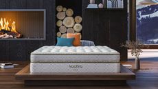2024's best mattress is the Saatva Classic luxury innerspring hybrid photographed here on a wooden bedframe in front of an open log fire and overlooking a snow-capped mountain
