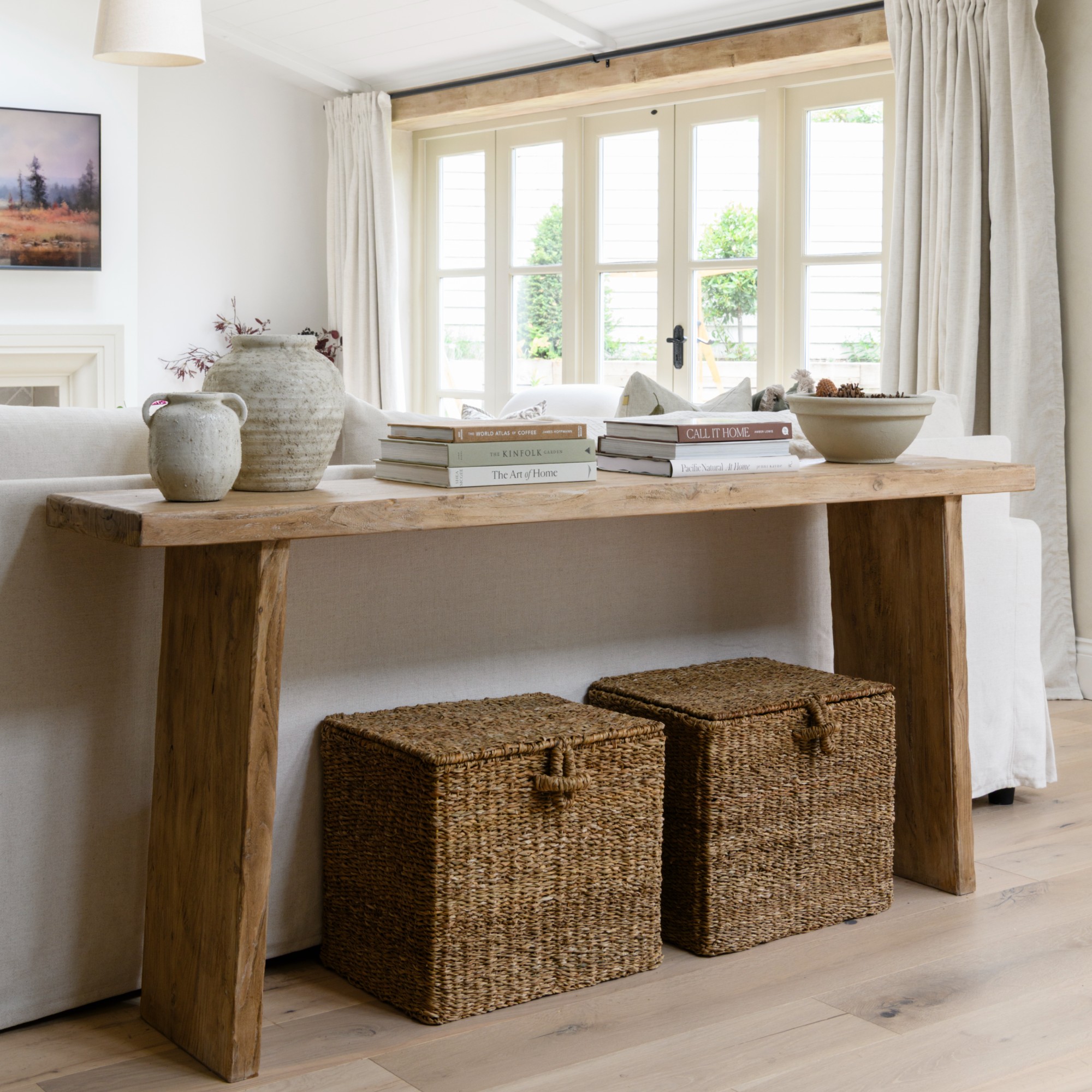 A white open-plan living room with a console table displaying coffee table books and ceramics and storage baskets underneath