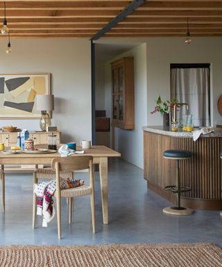 A modern kitchen with wooden beams, a wooden countertop with a bar stool, a light wooden dining table with decor on it and a painting in front of it, and a woven jute rug on the concrete floor