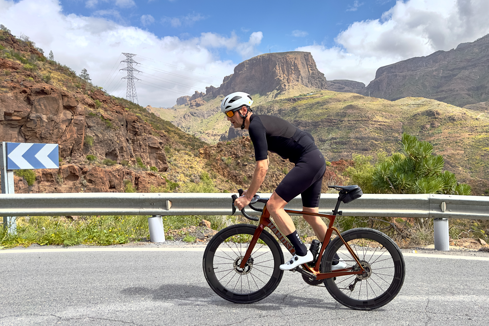 man riding an orange bike up a hill out of the saddle wearing black shorts, white helmet, white shoes and black jersey with hills in the background