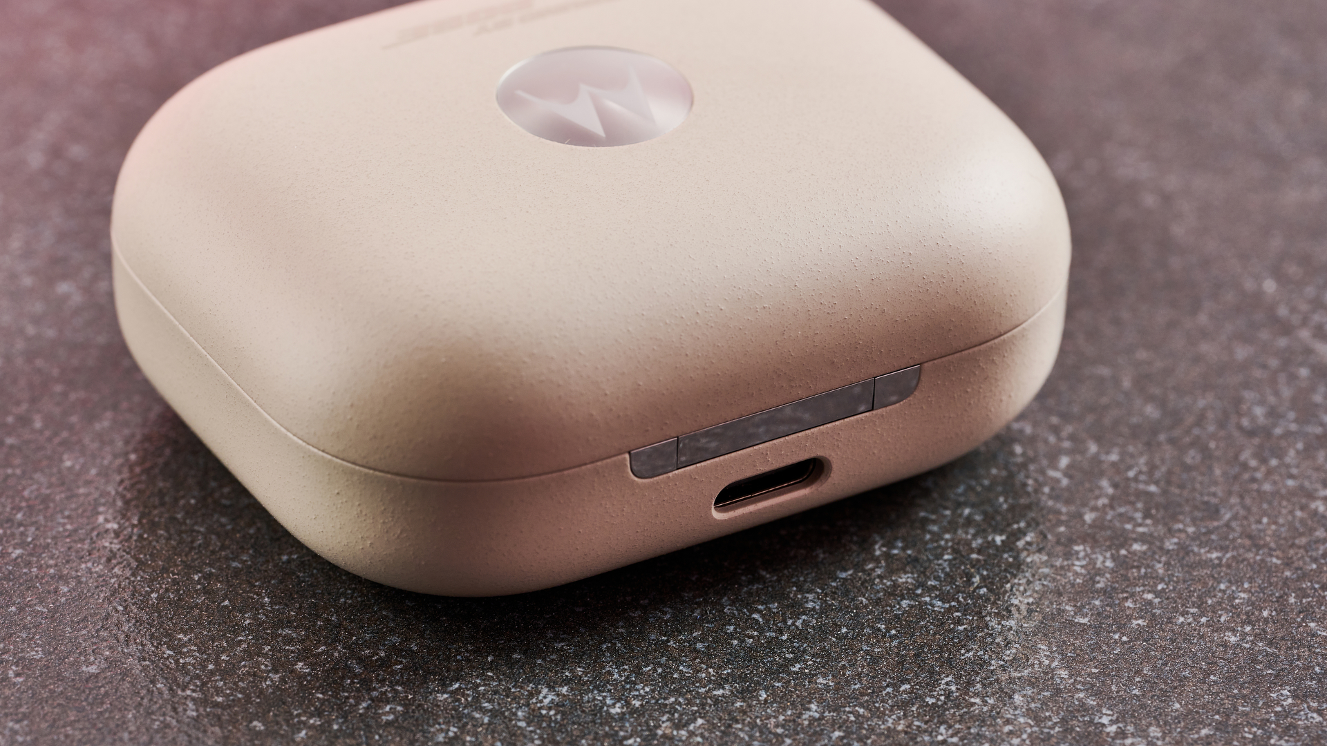 A close up of the front of the Motorola Moto Buds Plus case. It is closed and sitting on a dark surface and is against a pink background.