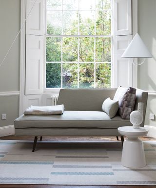 Gray daybed in front of tall white window, white low hanging pendant, geometric white stool, dark wooden flooring with patterned neutral rug