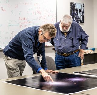 two men bending over images of galaxies. behind is a blackboard and an image of einstein