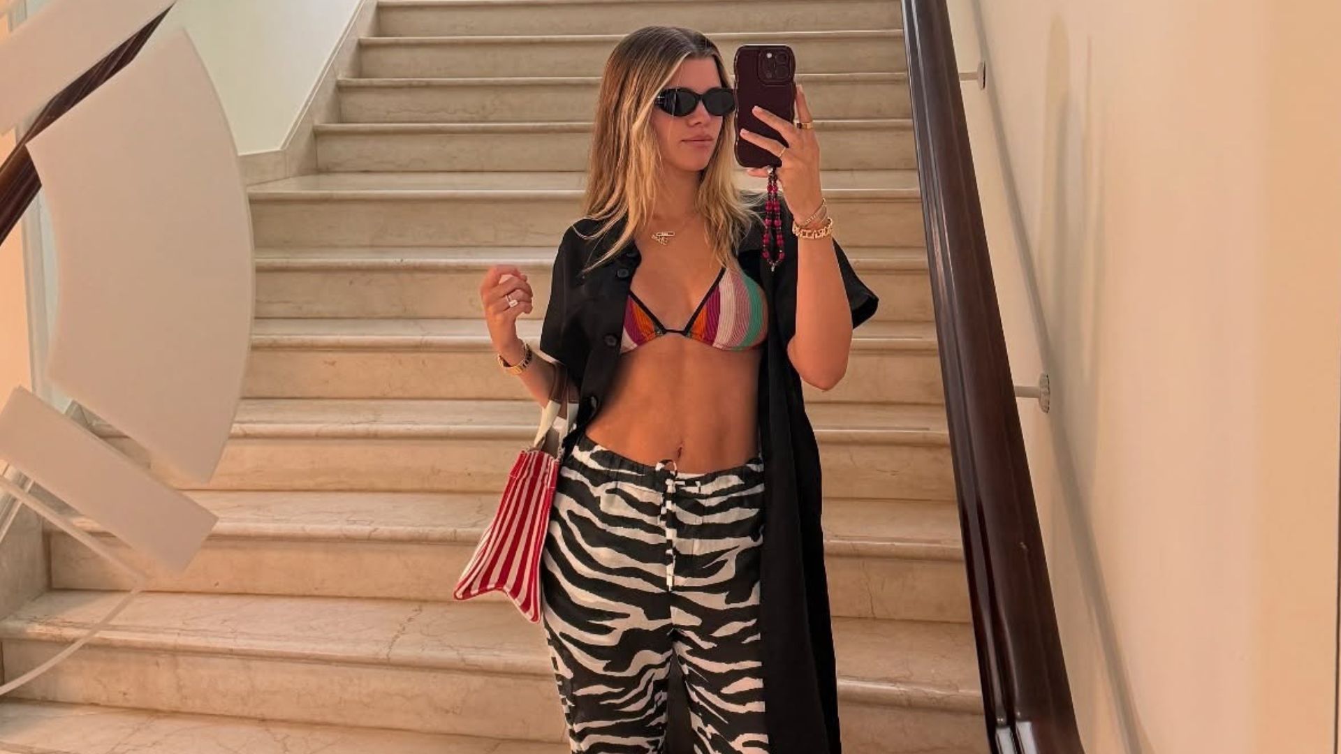 Sofia Richie Grainge stands at the bottom of a staircase taking a selfie in a mirror, wearing zebra print trousers with a multicoloured, striped bikini top and red flip-flops.