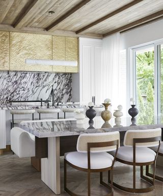 Dining with marble top dining table, dining chairs, and bench, bar and bar stools beyond and gold finish wall cabinets