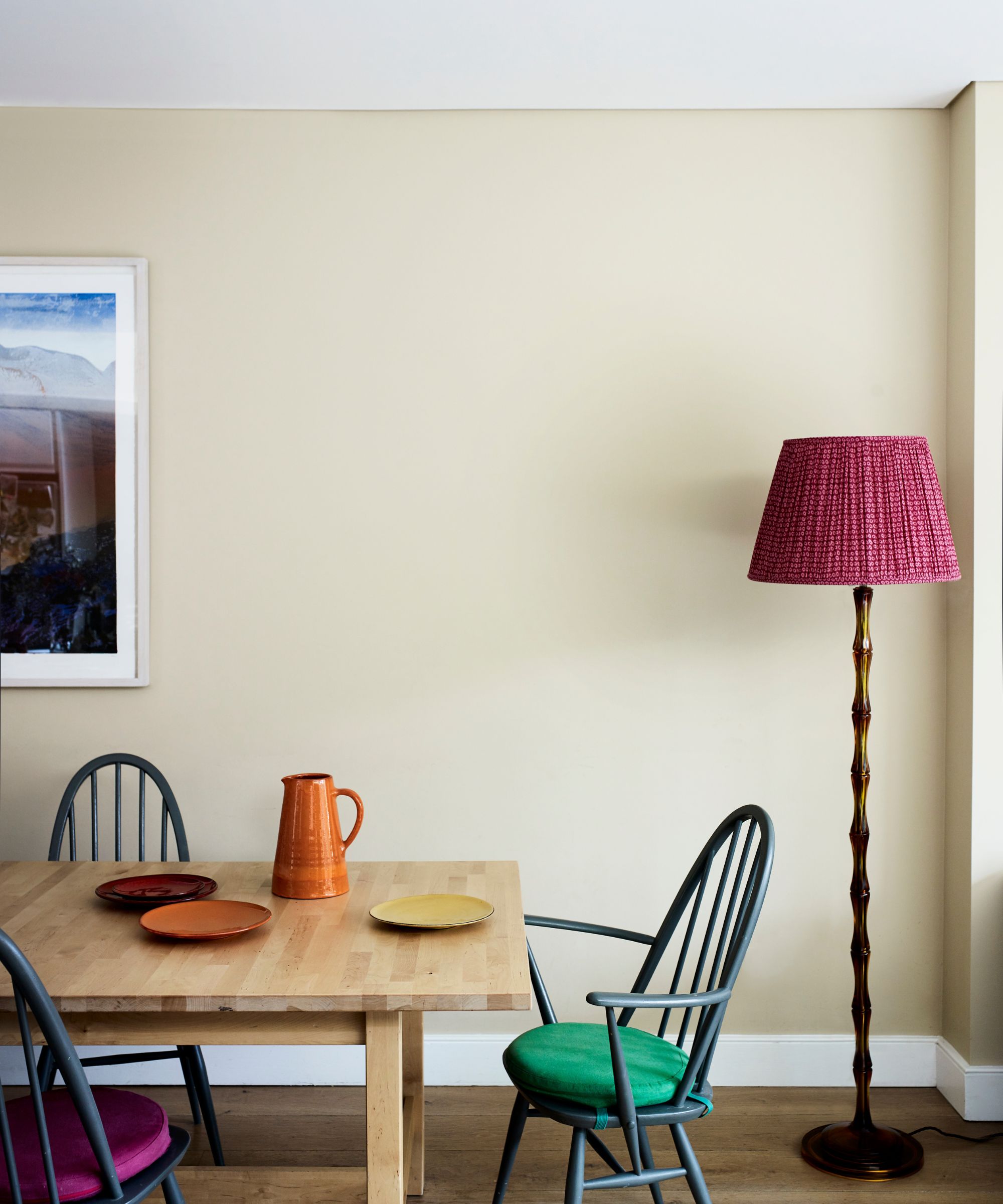 Dining room with magnolia walls, brass floor lamp with pink pleated shade in the corner, a wooden dining table with black wooden chairs and colourful cushions, and orange, red, and yellow tableware