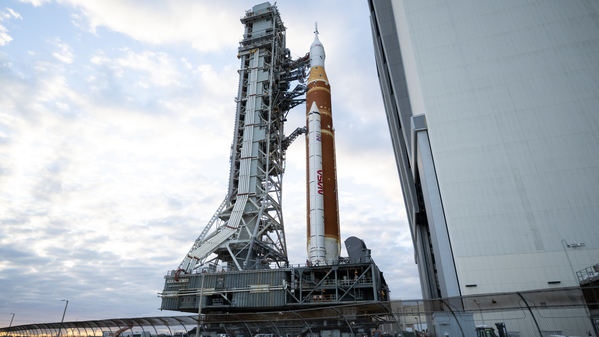 side view of a large orange and white rocket emerging from a huge gray building