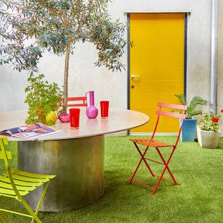 small garden with artificial lawn, bright chairs and accessories, table made around tree, yellow back door