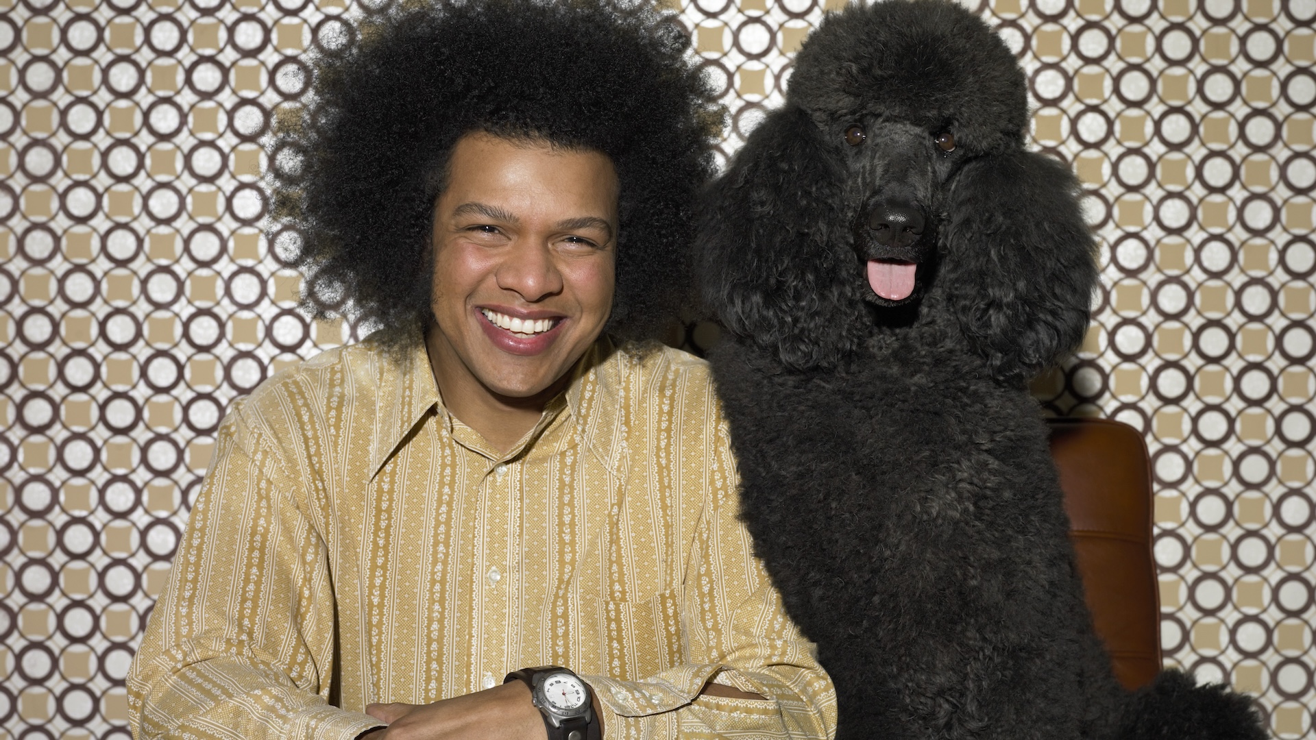 A man with a black afro sits next to his fluffy black poodle