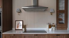 A section of a modern kitchen with wood panelling as the backsplash and a view of the extractor fan as well as the stovetop, cabinet and two wall lights hanging on either side