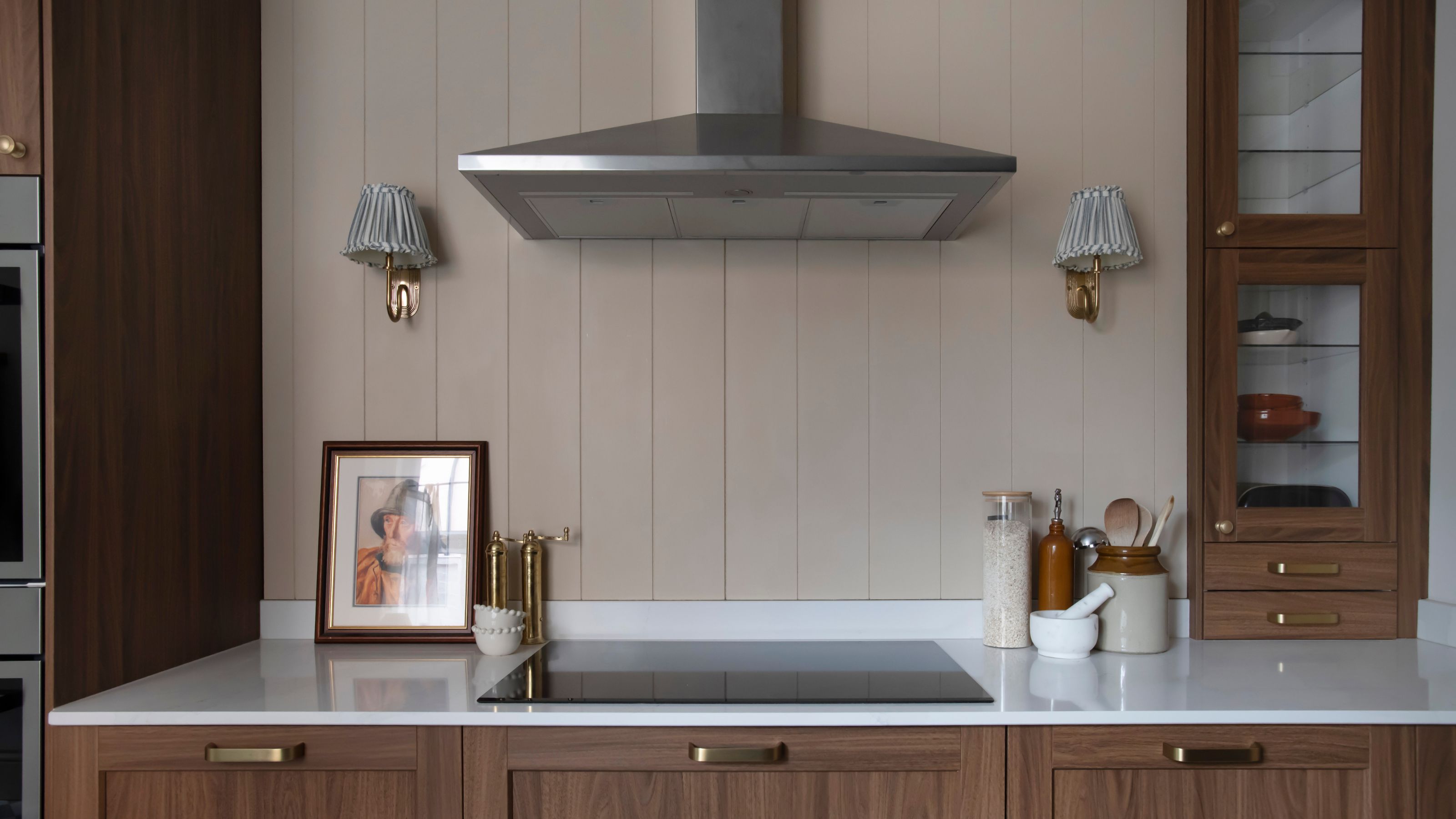 A section of a modern kitchen with wood panelling as the backsplash and a view of the extractor fan as well as the stovetop, cabinet and two wall lights hanging on either side 
