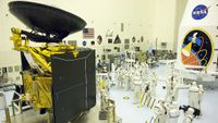 A large space probe stands in a big clean room full of people in white coveralls, across from a white rocket fairing with large mission logo.