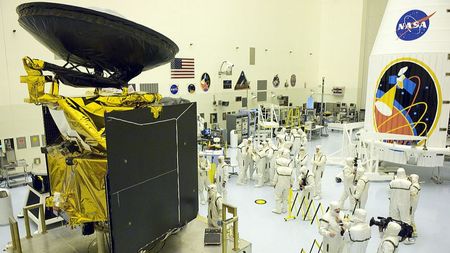 A large space probe stands in a big clean room full of people in white coveralls, across from a white rocket fairing with large mission logo.