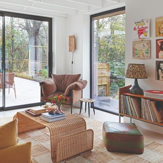 A 70s-style living room with large glass doors, a rattan coffee table and a table lamp and a wall light providing lighting