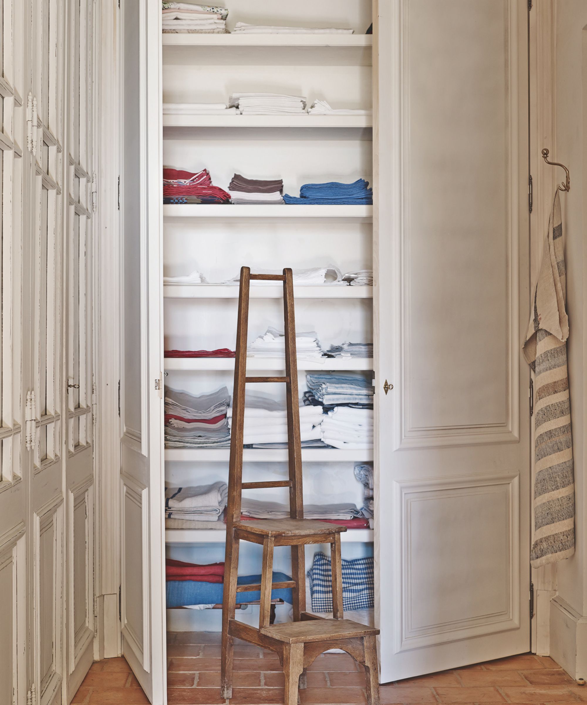 A white linen cupboard filled with folded laundry and a brown wooden ladder.
