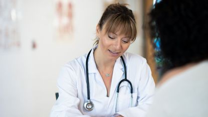 Woman conducting an NHS Health Check sat opposite woman with curly hair, back to camera