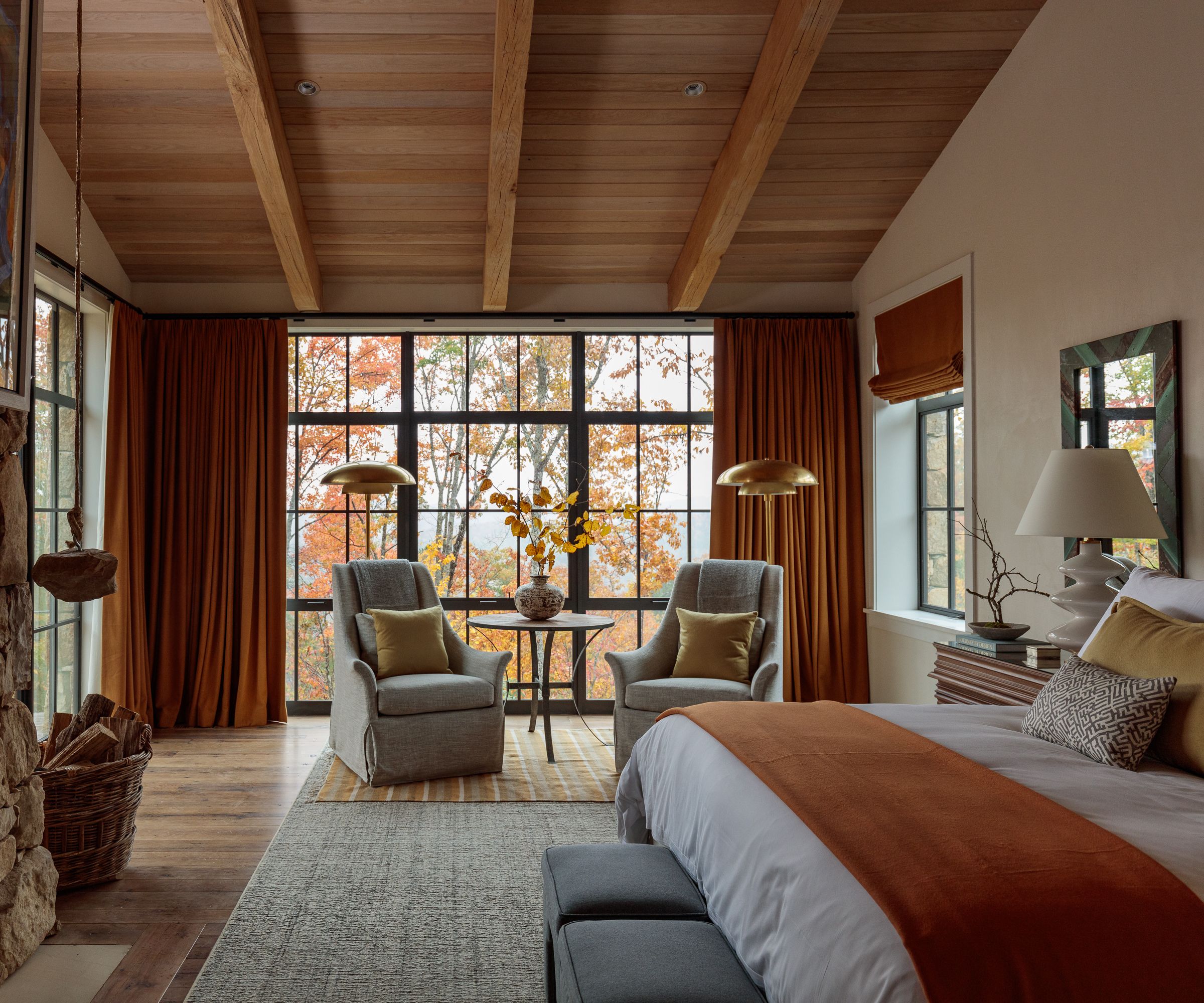 A neutral bedroom with a wooden sloped ceiling, large floor-to-ceiling windows and ochre curtains, and a throw on the bed.