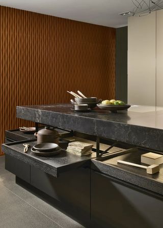 A dark colored kitchen island with pull out drawers and on the pull out drawers are plates and a cooking pot. On the island countertop is a bowl of fruit and a bowl with wooden spoons.