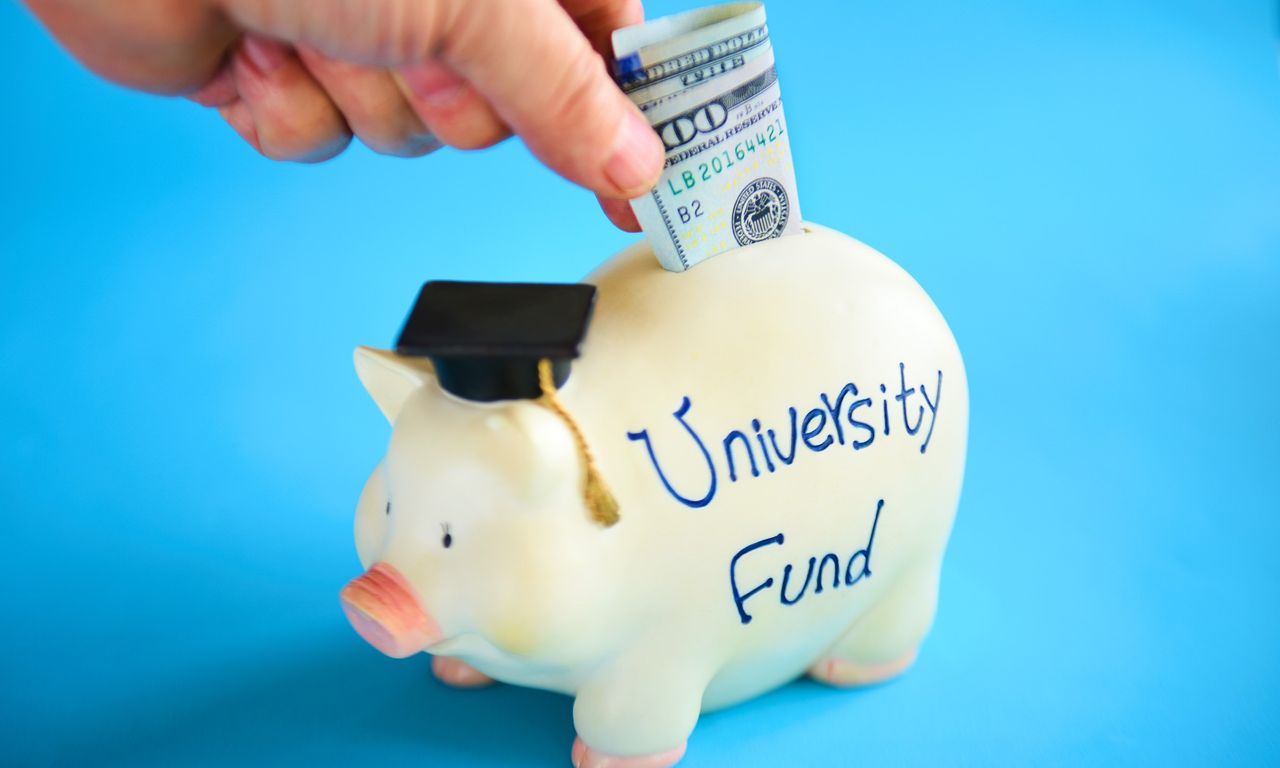 Hand putting a 100 dollar bill into a piggy bank that is wearing a graduation cap and has the words &quot;university fund&quot; written on its side