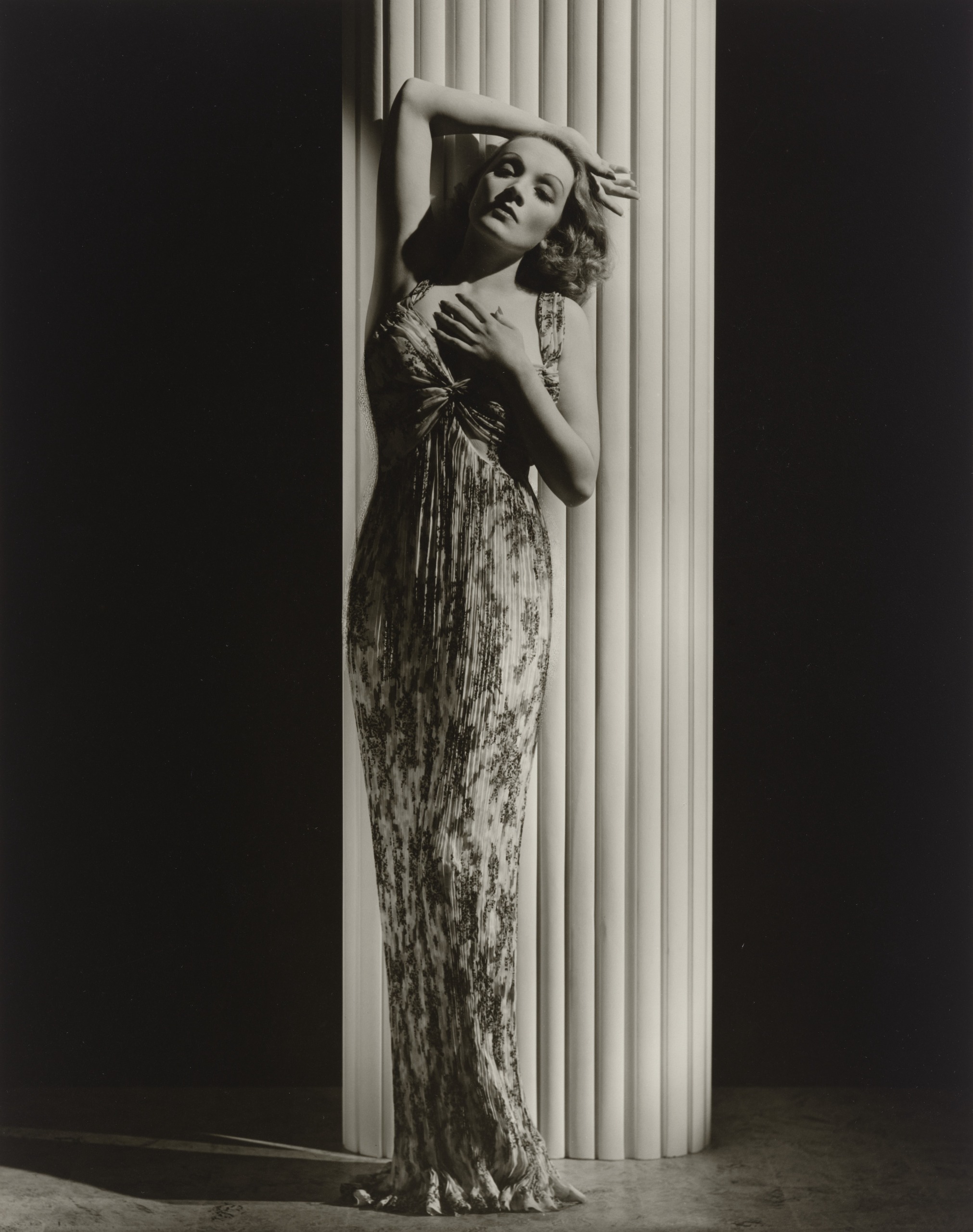 Marlene Dietrich in a long, pleated floral gown leans against a tall white fluted column with one arm raised over her head and a hand on her chest, her eyes half closed