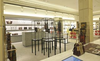 A Valentino store with various handbags displayed on wall shelves and metal framed glass cases with African masks displayed in them.