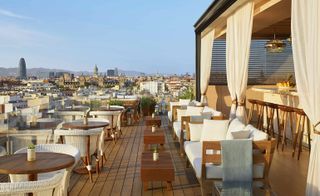 View of the rooftop dining space at The Edition Barcelona hotel, Spain featuring brown wooden tables, white seating, green plants and a bar area with stools and curtains. There is a view of nearby buildings under a blue sky