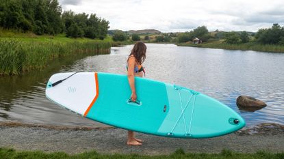 Woman holding Decathlon X100 SUP touring stand up paddle board next to a lake