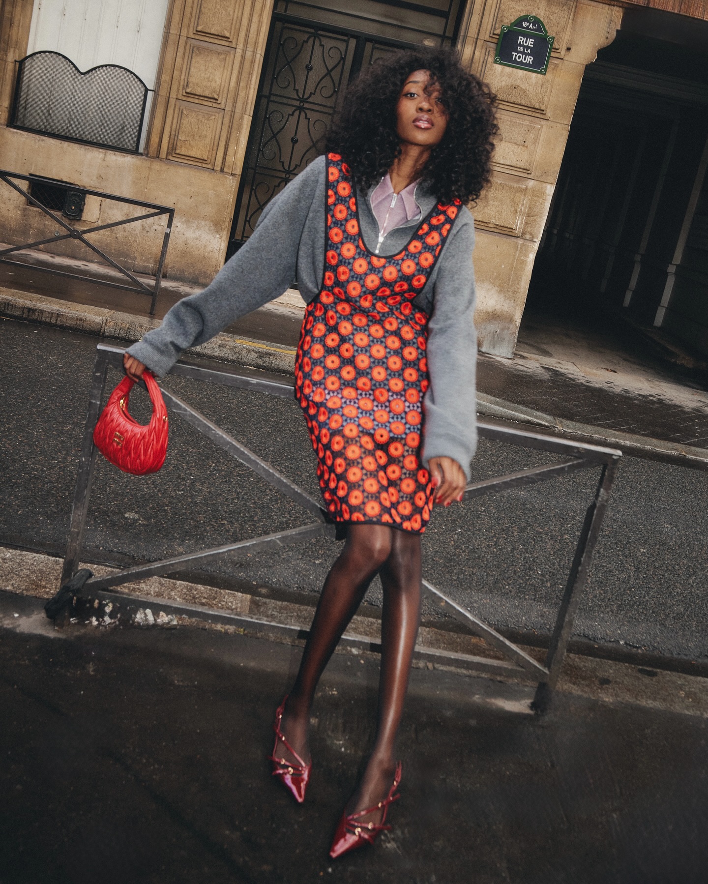 An image of an influencer wearing a red apron dress with a grey jumper underneath and holding a red Miu Miu bag and wearing red slingback heels