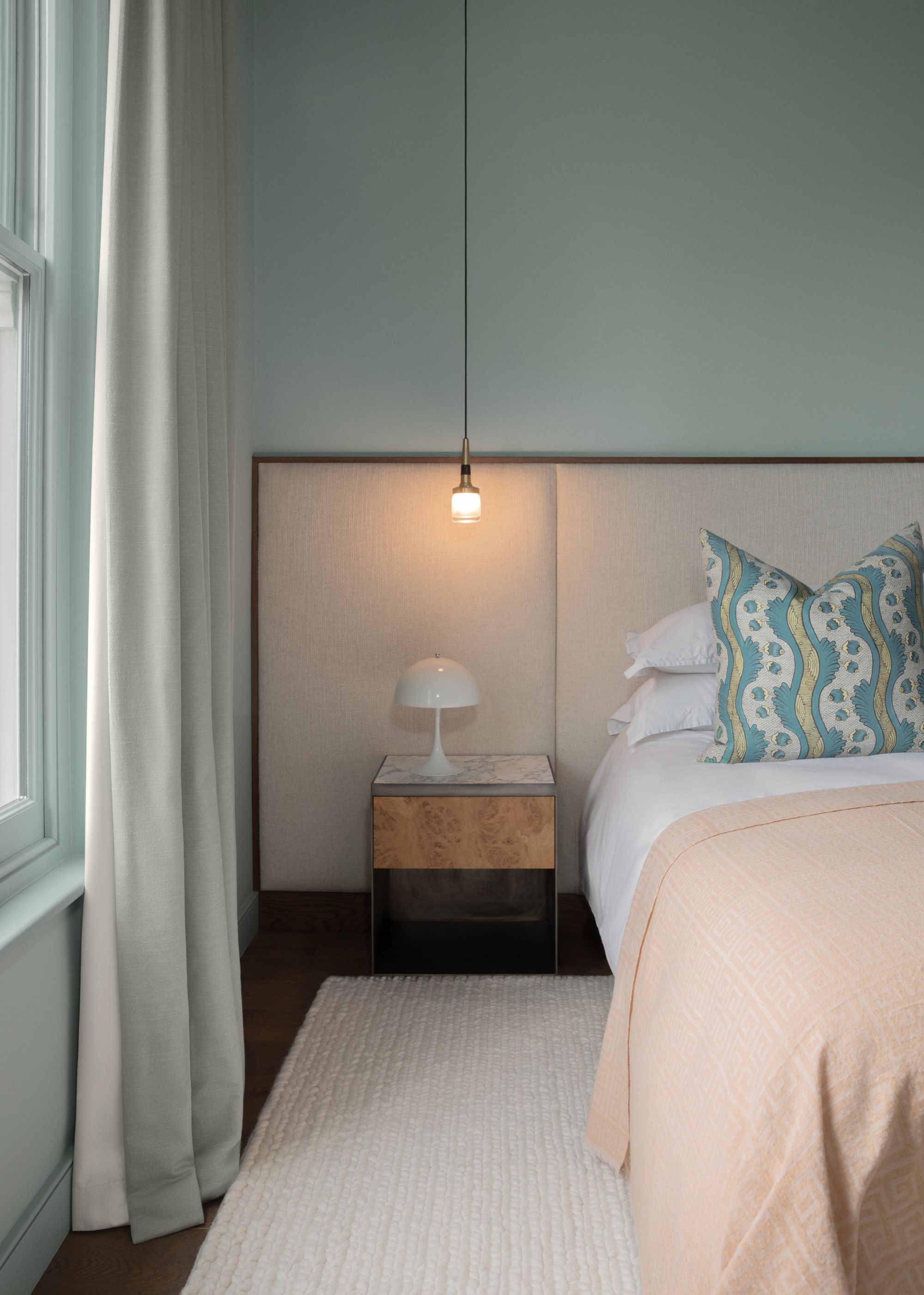 A bedroom with pale blue walls and white bedding with a pale pink throw. A beige rug and an oversized upholstered headboard.